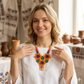 Femme blonde souriante portant une blouse blanche et un collier en perles aux couleurs vives (orange, rouge, bleu) dans un décor artisanal.