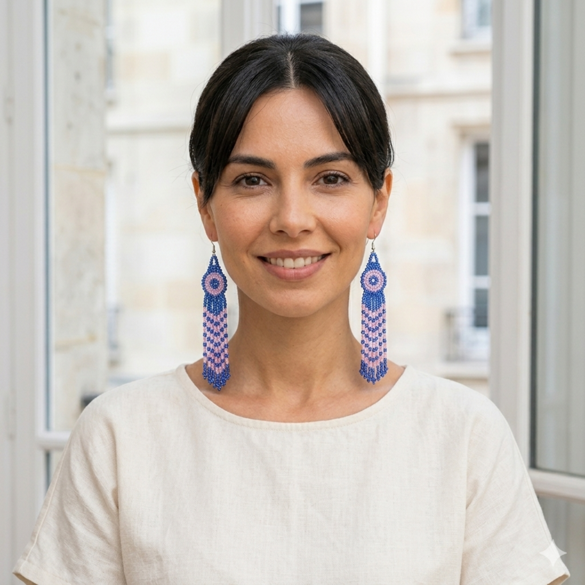 Portrait de face d'une femme souriante portant des boucles d'oreilles pendantes et longues en perles de rocaille bleues et roses tissées à la main, dans un style bohème chic.