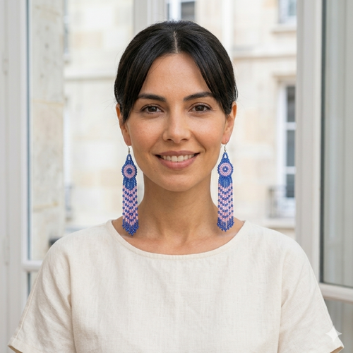 Portrait de face d'une femme souriante portant des boucles d'oreilles pendantes et longues en perles de rocaille bleues et roses tissées à la main, dans un style bohème chic.