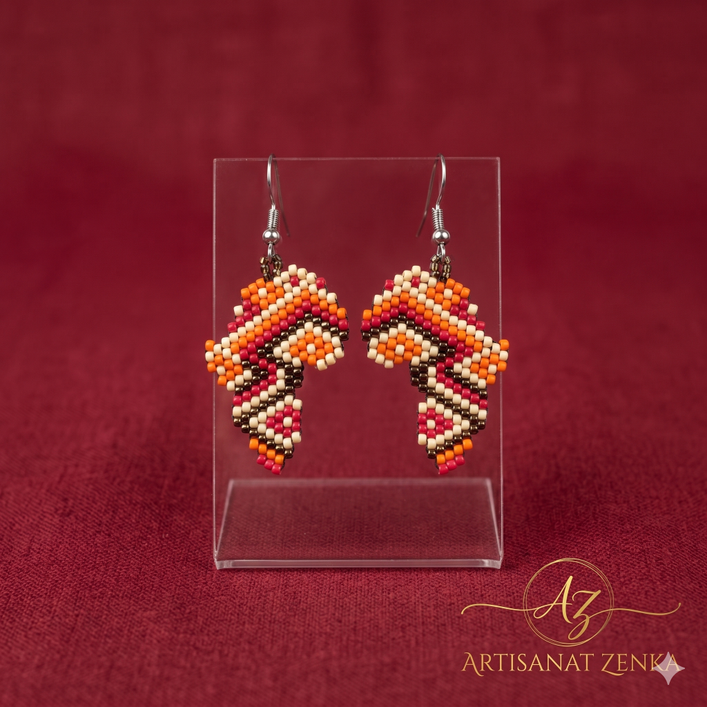 Vue de face de deux boucles d'oreilles en perles aux motifs géométriques orange et rouge sur un support transparent, fond texturé rouge grenade avec le logo doré Artisanat Zenka en bas à droite.