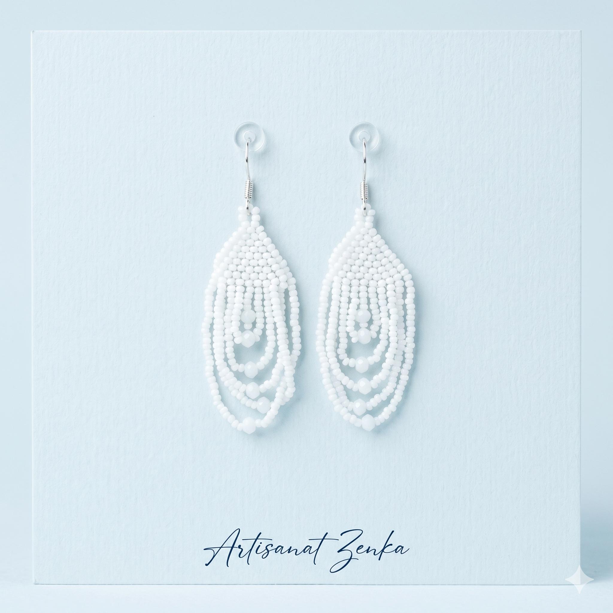 Paire de boucles d'oreilles pendantes en perles blanches de la marque Artisanat Zenka, présentées sur un support bleu pâle avec signature élégante.