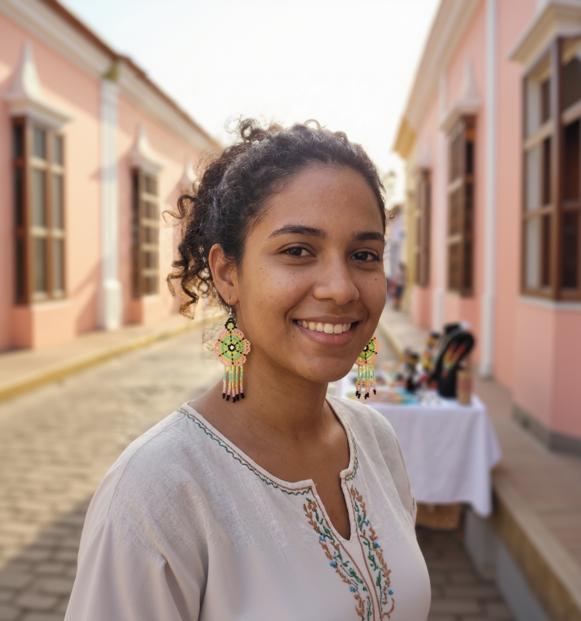 Portrait d'une femme souriante portant des boucles d'oreilles artisanales en perles multicolores (rose, vert, jaune) dans une rue ensoleillée.