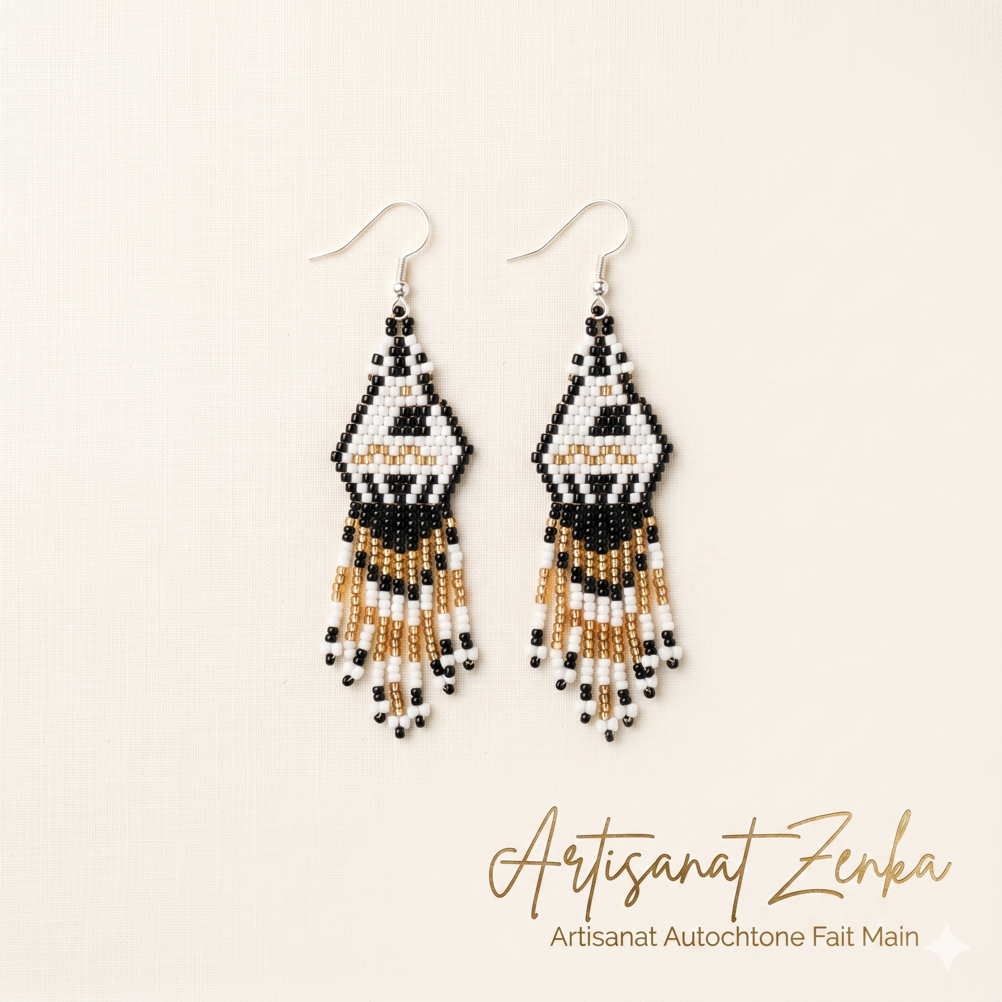 Paire de boucles d'oreilles artisanales en perles tissées avec motifs géométriques noirs et blancs et franges dorées sur fond crème, par Artisanat Zenka.