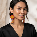 Portrait rapproché d'une femme souriante aux cheveux noirs attachés, portant de grandes boucles d'oreilles artisanales en perles de rocaille avec un motif solaire central rouge et jaune et des franges perlées. Elle porte un haut en satin noir élégant sur un fond texturé beige clair.