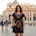Femme souriante portant une robe noire courte avec broderies florales colorées devant la Basilique Saint-Pierre à Rome, format carré pour boutique mode.