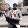Homme portant un t-shirt blanc "King" avec la carte de l'Afrique en motif ethnique coloré, assis sur une place à Londres.