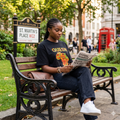 Femme élégante portant le t-shirt noir Queen Africa avec motif carte de l'Afrique en tissu coloré, assise sur un banc à Londres (St. Martin's Place)
