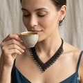 Gros plan d'une femme souriante buvant une tasse de café, portant un collier artisanal en perles noires et cristaux bleus assortis à ses boucles d'oreilles.