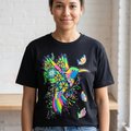 Portrait de face d'une femme souriante portant le t-shirt noir Les Ateliers Groupe BECCAR avec un motif graphique de colibri multicolore vibrant, dans un décor de studio lumineux.