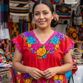Femme souriante portant une tunique rouge artisanale avec des broderies florales colorées devant un marché traditionnel.